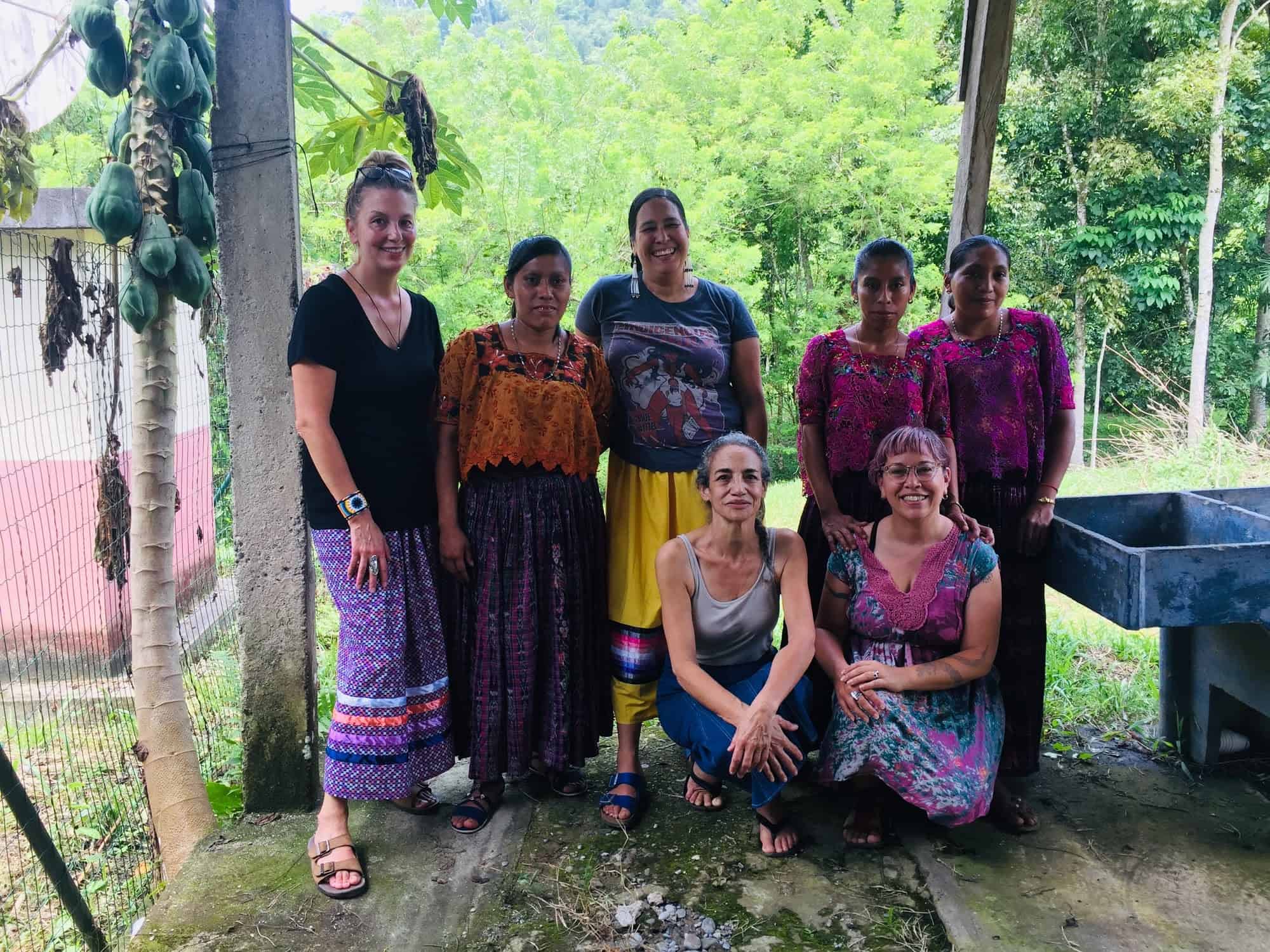 Seven women smile together, three in traditional Maya Q'eqchi' clothing. Bright green forest is behind them.