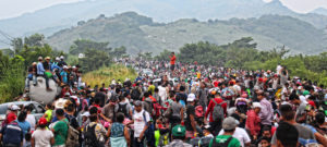 Asylum seekers fill the road while walking through Oaxaca, Mexico. In the background are rolling green hills. People are mostly on foot, and their clothing is bright and colorful in the foreground.