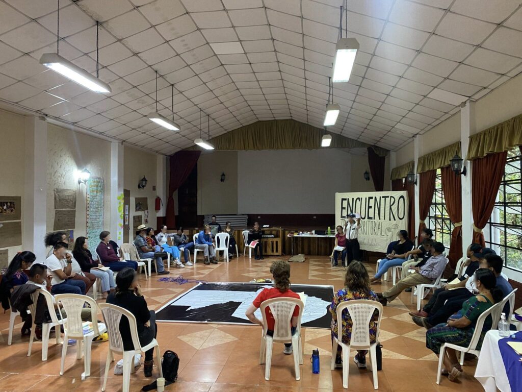 [ENG] In a room, there are people gathering sitting in chairs in a circle at the bottom of the room a white banner with black letter than said: "Encuentro Trans-Territorial" [ESP] En una sala, hay personas reunidas sentadas en sillas formando un circulo al fondo de la sala una pancarta blanca con letra negra que dice: "Encuentro Trans-Territorial"