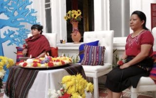 On the left corner there are yellow flowers and a cartoon blue tree. A woman dressed in red holds a microphone. In the center, there is a flower and candles offering. On the right another woman dressed in red and black, also holds a microphone and a red flower.