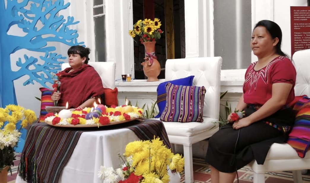 On the left corner there are yellow flowers and a cartoon blue tree. A woman dressed in red holds a microphone. In the center, there is a flower and candles offering. On the right another woman dressed in red and black, also holds a microphone and a red flower.