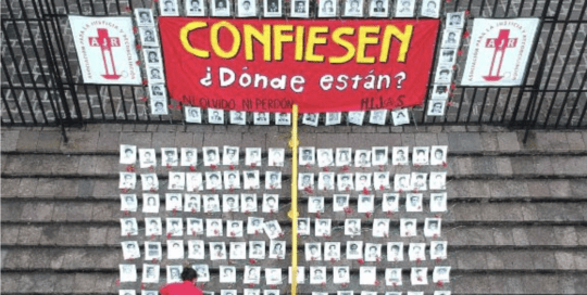 Alt text: Outside a fence there is a sign that reads “Confess: Where are they?” On the stairs, a person with a red t-shirt is laying the black and white pictures of the 183 disappeared who are registered in the Military Diary.
