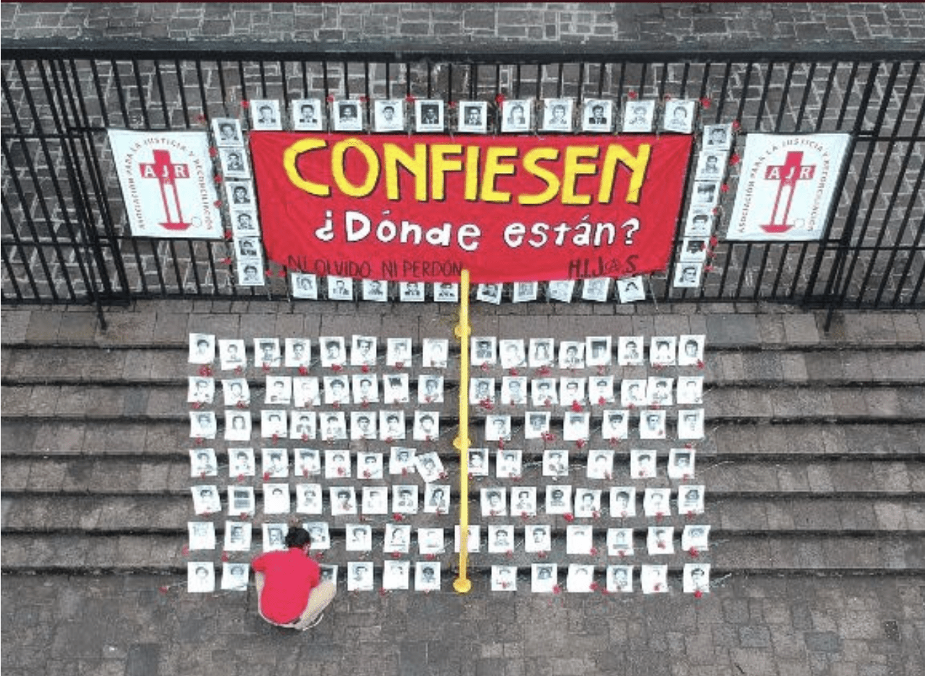 Alt text: Outside a fence there is a sign that reads “Confess: Where are they?” On the stairs, a person with a red t-shirt is laying the black and white pictures of the 183 disappeared who are registered in the Military Diary.