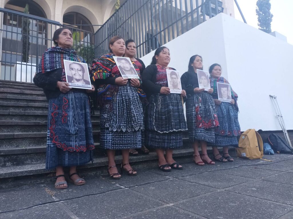 Six women in their traditional attire hold a photo in black and white of their relatives who were killed during the massacre on the Cumbre de Alaska case. Behind them are cement bleachers.
