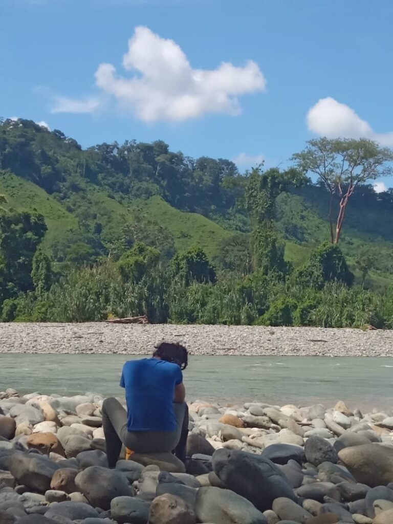 ESP: Una persona dando la espalda a la cámara, sentada sobre rocas a la orilla de un rio y grandes montañas verdes. ENG: 