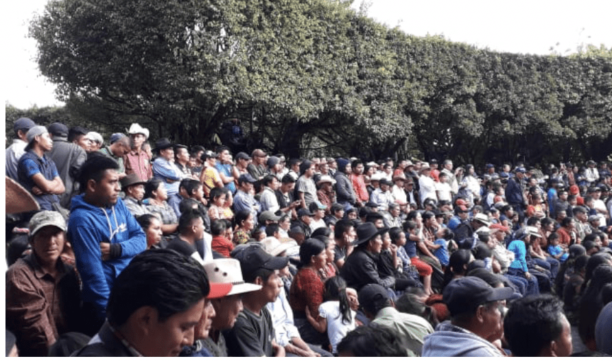 The picture shows a meeting of hundreds of people on Barillas waiting and hearing to someone not shown. Behind them there are trees and people are protecting themselves from the sun