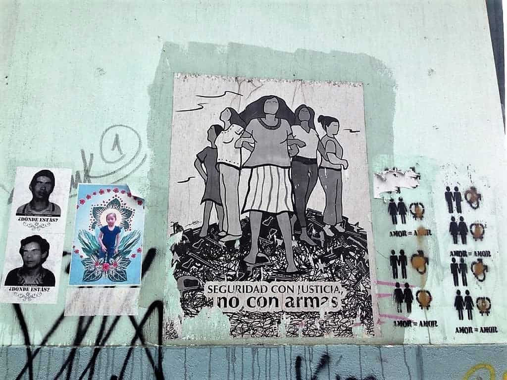 Street art in a wall at Guatemala City depicting in the center an image of women standing and protecting themselves with the legend "Security with justice not with weapons". In the left, pictures of disappeared during the Internal Armed conflict and of Jackelin Call who died under ICE custody. To the right a graffiti showing different couples equals love