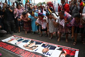 Grupo de Autoridades Indígenas con trajes tradicionales se reúnen en círculo alrededor de una lona extendida en el pavimento que muestra fotografías de funcionaries públiques. Las mujeres sostienen bastones ceremoniales mientras participantes y medios de comunicación rodean la escena.