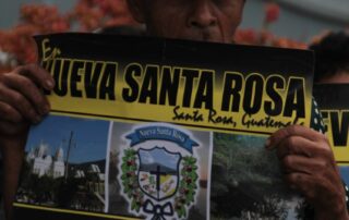 There is a man with a beige cowboy hat, the man holds with both hands a poster that has pictures of nature, and says: "In Nueva Santa Rosa, Santa Rosa, Guatemala. 98.86% said: NO TO MINING.