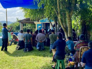 People gathered outdoors in front of a small blue monument or altar, some standing and others kneeling, participating in a ceremony under the shade of trees. Men and women of various ages are present, including some wearing traditional clothing. The scene is calm and communal./Personas reunidas al aire libre frente a un pequeño monumento o altar azul, algunas de pie y otras de rodillas, participando en una ceremonia bajo la sombra de árboles. Hay hombres y mujeres de distintas edades, algunos usando trajes tradicionales. El ambiente es tranquilo y comunitario.