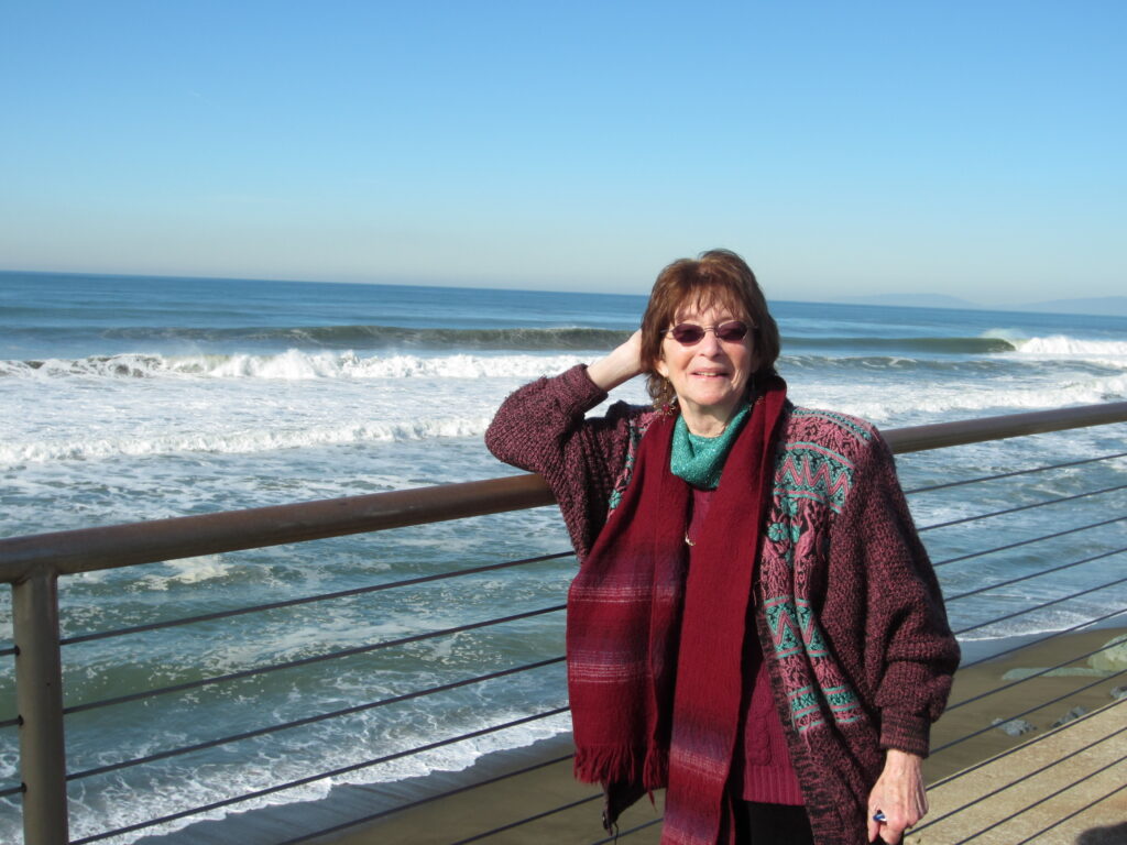 In the back there is the ocean and a bridge, Susan Jones, wearing sunglasses and a red coat stands in the bridge while looking at front