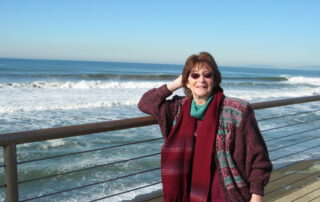In the back there is the ocean and a bridge, Susan Jones, wearing sunglasses and a red coat stands in the bridge while looking at front