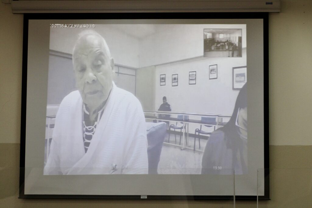 Thursday, July 25, Hearing 55 The photo shows an adult male in a bathrobe on a screen looking straight ahead. A women can be seen partially at the left