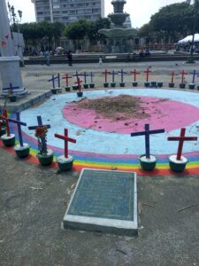 There are two concentric circles painted on the ground, first pink then blue. Along the ring there is a rainbow painted on the ground and on top of the rainbow rests 41 crosses to conmemorate the girls who lost their lives in the Safe House fire. There is an official plaque in the forefront. The photo is taken outside in Guatemala City's central plaza.