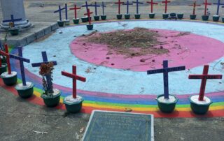 There are two concentric circles painted on the ground, first pink then blue. Along the ring there is a rainbow painted on the ground and on top of the rainbow rests 41 crosses to conmemorate the girls who lost their lives in the Safe House fire. There is an official plaque in the forefront. The photo is taken outside in Guatemala City's central plaza.