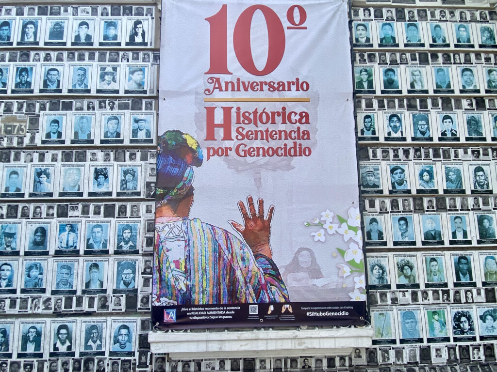 Behind a wallpaper of faces of the disappeared in the house of memory, hangs a banner on which in red letters reads the tenth anniversary of the historic sentence for genocide. Below is the back of an Ixil woman with her hand raised giving her testimony before Guatemalan courts.