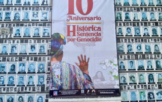 Behind a wallpaper of faces of the disappeared in the house of memory, hangs a banner on which in red letters reads the tenth anniversary of the historic sentence for genocide. Below is the back of an Ixil woman with her hand raised giving her testimony before Guatemalan courts.
