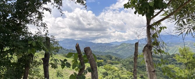 Vista de un paisaje montañoso enmarcado por árboles y una cerca rústica de madera en primer plano. El cielo está parcialmente nublado y el valle abajo está cubierto de vegetación verde.