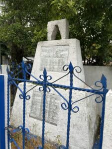 White concrete monument with engraved plaques behind a blue wrought-iron fence. Trees are visible around the monument.