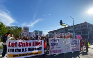 In a street, a group of people carry banners with messages about the People's Summit for Democracy, about the US economic blockade of Cuba, and more.