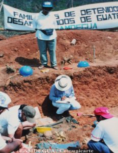 FAMDEGUA is supervising and exhumation on Los Josefinos Village, there are three people on the ground searching for bones and one person standing in front of a sign that says FAMDEGUA