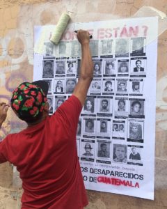 A man with a red t-shirt and a hat is posting a sign with faces of the disappeared. On top on red the sign reads Where are they?