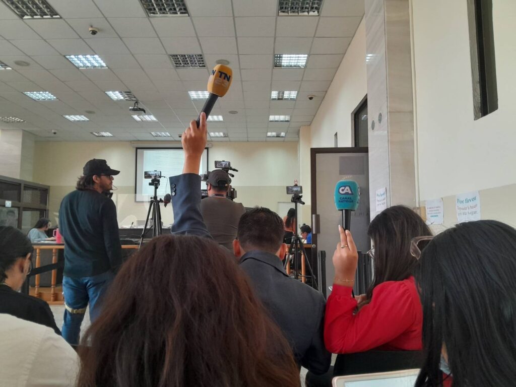 in first plane in the court room people sitting and journalist have their microphones up to record and there is a person stading doing video recording