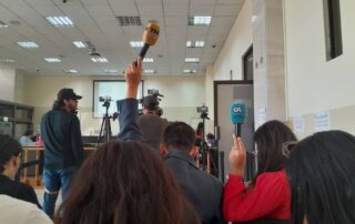 in first plane in the court room people sitting and journalist have their microphones up to record and there is a person stading doing video recording