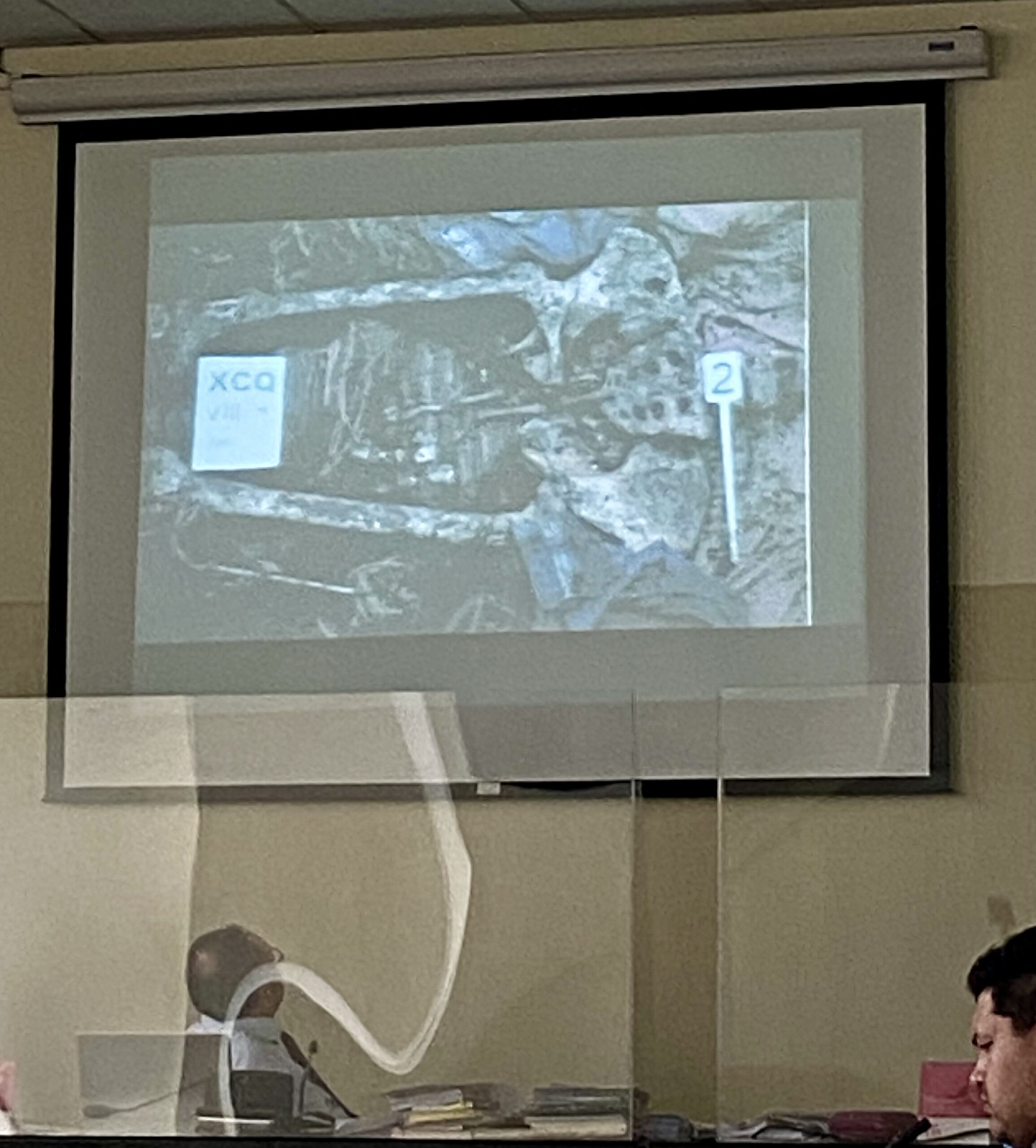 The photo above the court screen shows a photo of one of the graves where a skeleton can be seen. Below is the judge looking at the photo and on the left is the face of a man sitting in the row of lawyers. ////en foto se observa arriba la pantalla del tribunal se observa una foto de uno de las fosas donde se puede ver una osamenta. Abajo se observa al juez titular mirando la fotografia y se observa en la izquierda el rostro de un hombre sentado en la fila de abogados