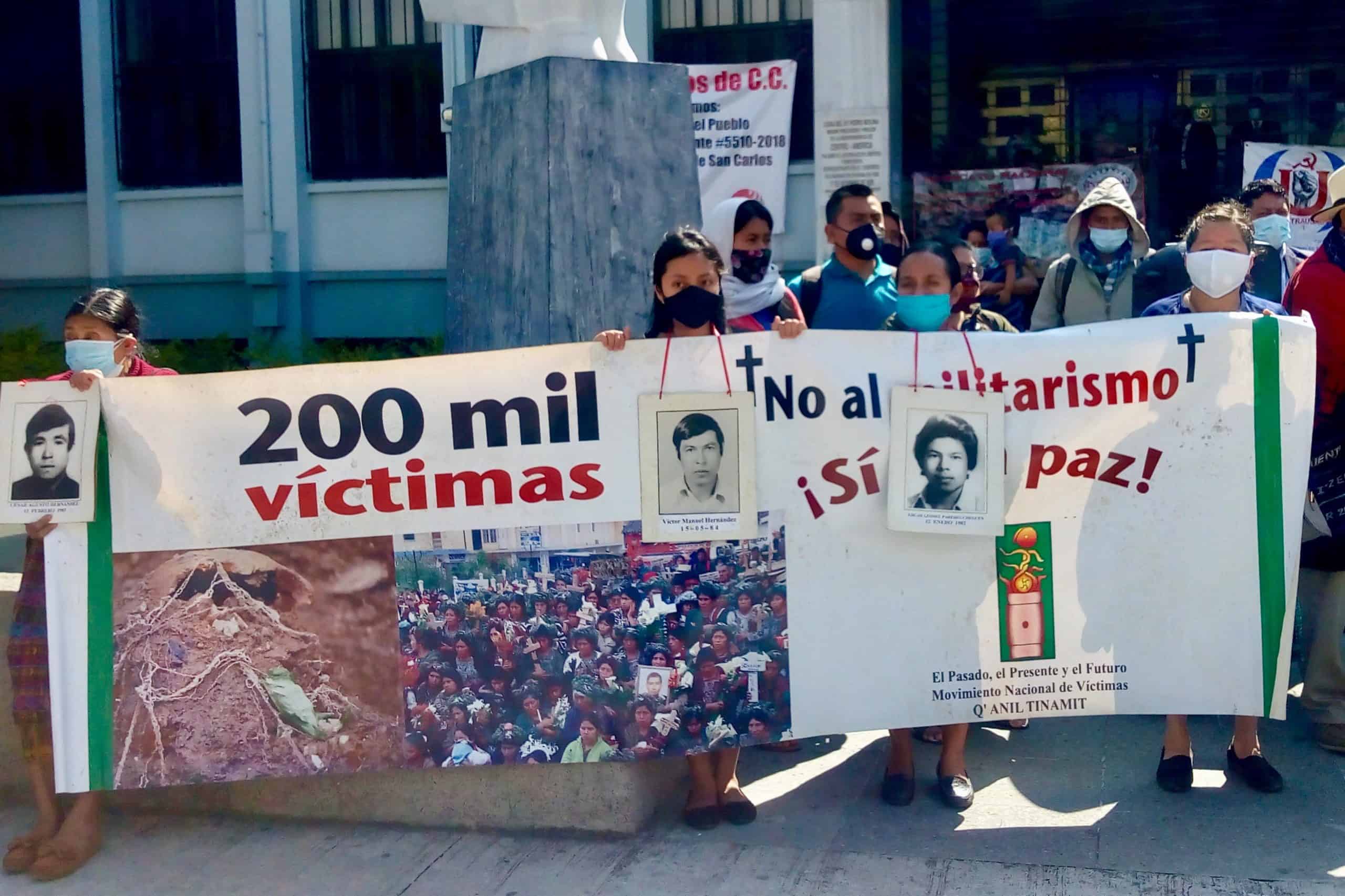 On the left a woman holds both a picture of a disappeared love one and a sign that reads 200, 000 victims. On the other side, there are a group of 10 people behind a sign that reads No to militarism, yes to Peace! All of them are wearing masks. 