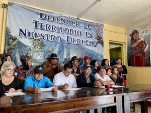 Fifteen people sit and stand together during a press conference. Behind them, a large banner reads 'Defending the Land is Our Right' in Spanish. Some individuals are holding the traditional vara of Indigenous authority. A large portrait of an Indigenous woman in traditional dress is displayed on the right wall.