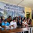 Fifteen people sit and stand together during a press conference. Behind them, a large banner reads 'Defending the Land is Our Right' in Spanish. Some individuals are holding the traditional vara of Indigenous authority. A large portrait of an Indigenous woman in traditional dress is displayed on the right wall.