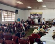 A large group of people sits in a community hall, facing a stage where several individuals stand and speak. The audience includes many women wearing traditional Ixil clothing and woven headdresses. On the stage, a marimba band is set up to the right, and a banner related to genocide justice is displayed on the wall. Tables are arranged in rows, and a ceremonial offering is placed on the floor in front of the stage. Bright daylight filters through the large windows on the left side of the room.