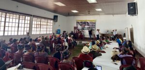 A large group of people sits in a community hall, facing a stage where several individuals stand and speak. The audience includes many women wearing traditional Ixil clothing and woven headdresses. On the stage, a marimba band is set up to the right, and a banner related to genocide justice is displayed on the wall. Tables are arranged in rows, and a ceremonial offering is placed on the floor in front of the stage. Bright daylight filters through the large windows on the left side of the room.