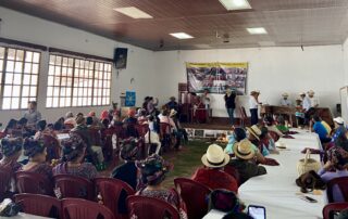 A large group of people sits in a community hall, facing a stage where several individuals stand and speak. The audience includes many women wearing traditional Ixil clothing and woven headdresses. On the stage, a marimba band is set up to the right, and a banner related to genocide justice is displayed on the wall. Tables are arranged in rows, and a ceremonial offering is placed on the floor in front of the stage. Bright daylight filters through the large windows on the left side of the room.