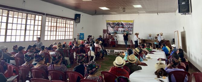 A large group of people sits in a community hall, facing a stage where several individuals stand and speak. The audience includes many women wearing traditional Ixil clothing and woven headdresses. On the stage, a marimba band is set up to the right, and a banner related to genocide justice is displayed on the wall. Tables are arranged in rows, and a ceremonial offering is placed on the floor in front of the stage. Bright daylight filters through the large windows on the left side of the room.