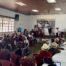 A large group of people sits in a community hall, facing a stage where several individuals stand and speak. The audience includes many women wearing traditional Ixil clothing and woven headdresses. On the stage, a marimba band is set up to the right, and a banner related to genocide justice is displayed on the wall. Tables are arranged in rows, and a ceremonial offering is placed on the floor in front of the stage. Bright daylight filters through the large windows on the left side of the room.