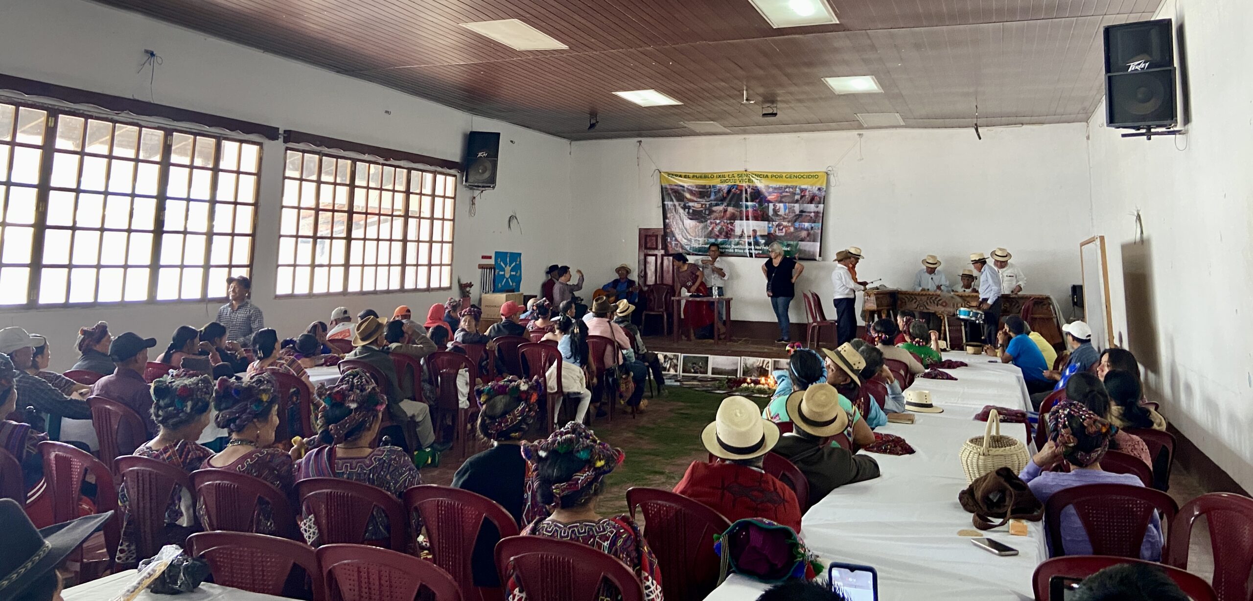 A large group of people sits in a community hall, facing a stage where several individuals stand and speak. The audience includes many women wearing traditional Ixil clothing and woven headdresses. On the stage, a marimba band is set up to the right, and a banner related to genocide justice is displayed on the wall. Tables are arranged in rows, and a ceremonial offering is placed on the floor in front of the stage. Bright daylight filters through the large windows on the left side of the room.