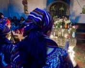 A group of Indigenous women wearing colorful woven garments and striped headscarves participates in a traditional Maya ceremony. In front of them is an altar decorated with lit candles, flowers in vases, and a small fire on the ground. The ceremony takes place indoors, with other people seated in the background and symbolic artwork painted on the walls.