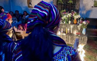 A group of Indigenous women wearing colorful woven garments and striped headscarves participates in a traditional Maya ceremony. In front of them is an altar decorated with lit candles, flowers in vases, and a small fire on the ground. The ceremony takes place indoors, with other people seated in the background and symbolic artwork painted on the walls.