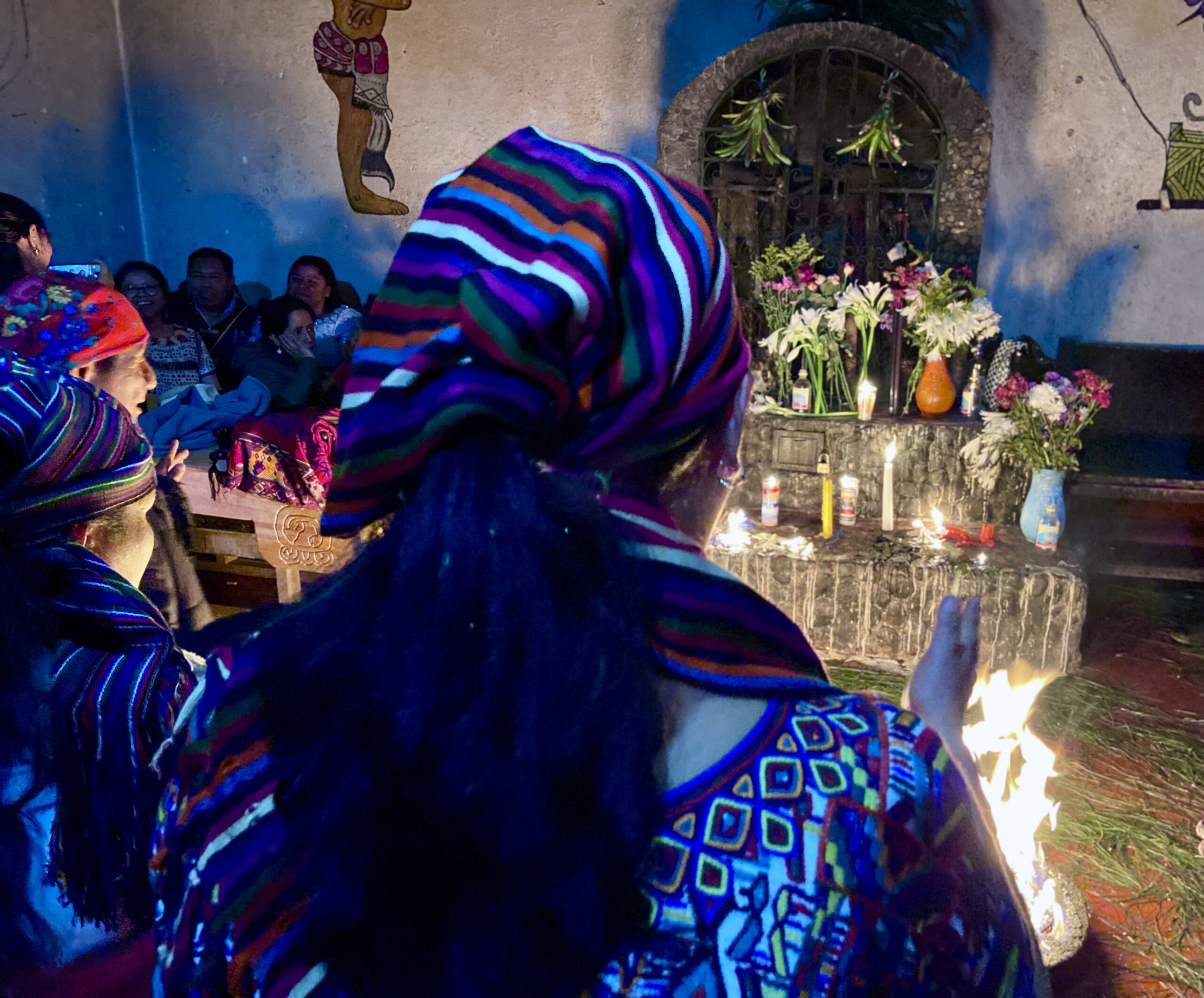 A group of Indigenous women wearing colorful woven garments and striped headscarves participates in a traditional Maya ceremony. In front of them is an altar decorated with lit candles, flowers in vases, and a small fire on the ground. The ceremony takes place indoors, with other people seated in the background and symbolic artwork painted on the walls.