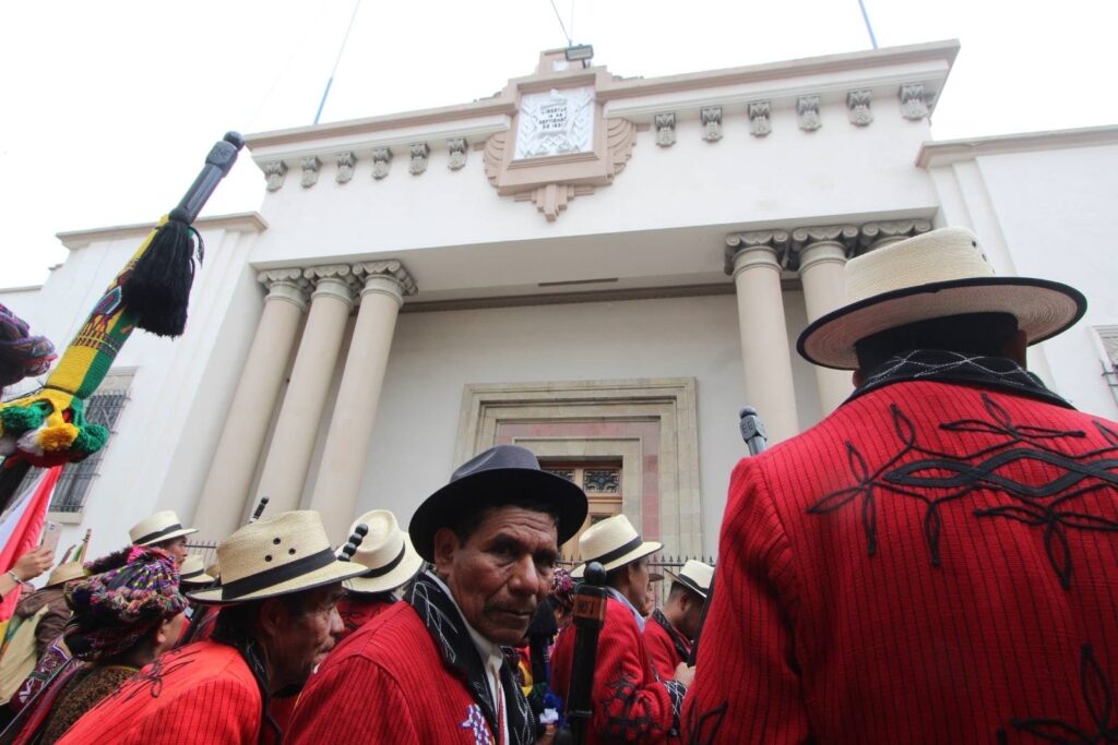 Frente al Ministerio Público un edificio beige un grupo de Autoridades Indígenas con su traje tradicional en color rojo, sombrero beige y su bastón sagrado.