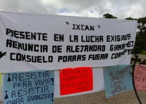 In the middle of a road, you can see clouds in the background, there is a white blanket that says Ixcán present in the fight, we demand the resignation of Alejandro Giammattei and Consuelo Porras. Below on the left a blue sign that says Resist to Live, Resist to Advance. Next, an orange sign that says resign Giammattei! And Porras Out! on the right side a sign says the people united will never be defeated.