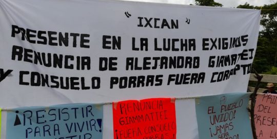 In the middle of a road, you can see clouds in the background, there is a white blanket that says Ixcán present in the fight, we demand the resignation of Alejandro Giammattei and Consuelo Porras. Below on the left a blue sign that says Resist to Live, Resist to Advance. Next, an orange sign that says resign Giammattei! And Porras Out! on the right side a sign says the people united will never be defeated.