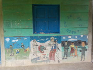 A painting of a school room. The upper wall has green paint with blue windows and in the below there is a mural painted by children depicting scenes of the attacks on Indigenous Communities during the Internal Armed Conflict