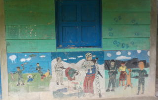 A painting of a school room. The upper wall has green paint with blue windows and in the below there is a mural painted by children depicting scenes of the attacks on Indigenous Communities during the Internal Armed Conflict