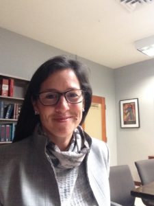 Photograph of Lydia Lopez smiling in an office. She is femme with long dark hair and is wearing a grey suit.