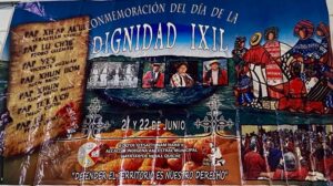 A large, colorful banner hung on a tiled ceiling, announcing the "Commemoration of the Day of Ixil Dignity." On the left side, a scroll-like graphic lists the names of the Seven Principales—Sebastián Cedillo, Pedro Guzmán, Vicente Guzmán, Juan Brito, Juan Brito Brito, Diego Cuchil, and Pedro Saloj—who were executed in 1936 for resisting forced labor. In the center and right, the banner features a collage of photographs: black-and-white images of community leaders, color portraits of people in traditional Ixil clothing, and stylized Indigenous artwork showing a group of figures in red, blue, and white garments. At the bottom, there are more event photos, maize kernels as background, and the phrase: “Defender el territorio es nuestro derecho” ("Defending the land is our right"). The banner credits the Ancestral Indigenous Municipal Authority Maya Ixil of Nebaj, Quiché, with organizing the event, held on June 21 and 22.