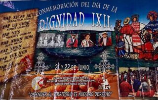 A large, colorful banner hung on a tiled ceiling, announcing the "Commemoration of the Day of Ixil Dignity." On the left side, a scroll-like graphic lists the names of the Seven Principales—Sebastián Cedillo, Pedro Guzmán, Vicente Guzmán, Juan Brito, Juan Brito Brito, Diego Cuchil, and Pedro Saloj—who were executed in 1936 for resisting forced labor. In the center and right, the banner features a collage of photographs: black-and-white images of community leaders, color portraits of people in traditional Ixil clothing, and stylized Indigenous artwork showing a group of figures in red, blue, and white garments. At the bottom, there are more event photos, maize kernels as background, and the phrase: “Defender el territorio es nuestro derecho” ("Defending the land is our right"). The banner credits the Ancestral Indigenous Municipal Authority Maya Ixil of Nebaj, Quiché, with organizing the event, held on June 21 and 22.
