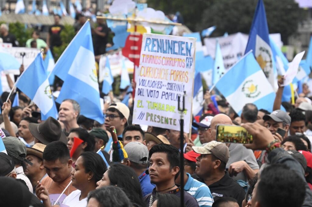 Una manifestación, alzan banderas de guatemala, bastones ancestrales y se lee un cartel que dice: “Renuncien tiranos corruptos, violadores de soberanía del voto popular”